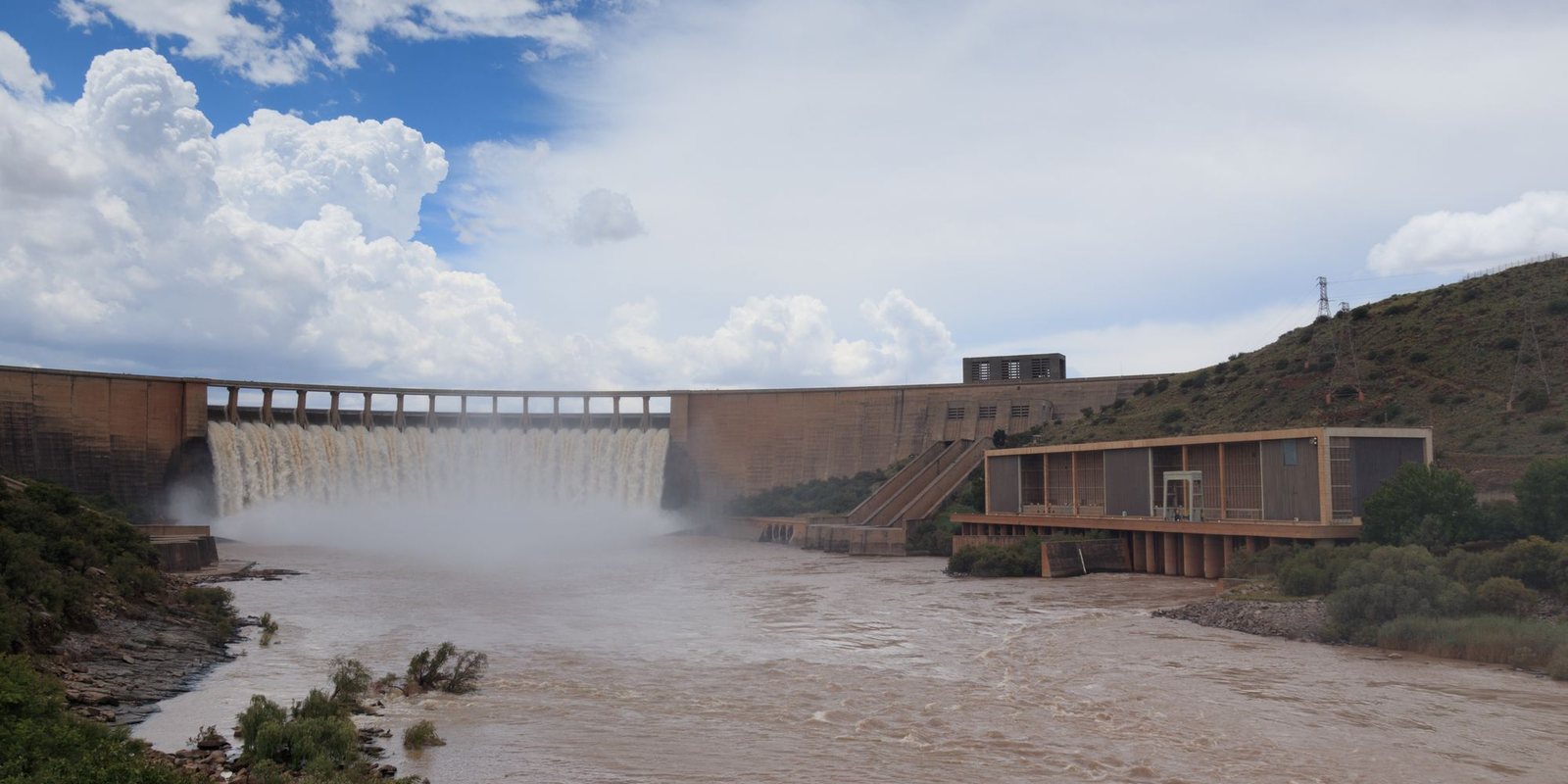 Barragem com vertedouro aberto, casa de força à direita e nuvens carregadas sobre o vale