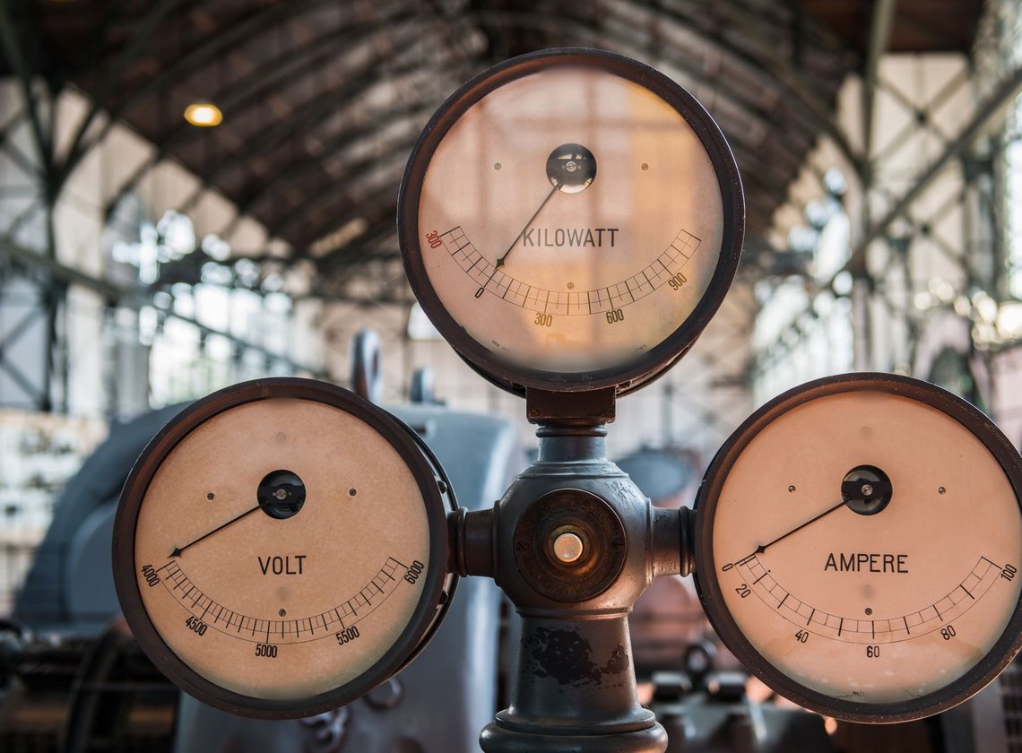 Três mostradores antigos de quilowatt, volt e ampere montados num mesmo suporte dentro de um galpão industrial