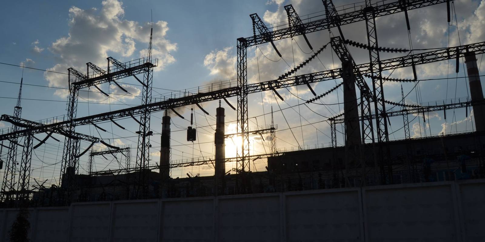 Estruturas metálicas e cabos de uma subestação de transformação em silhueta, com o sol atrás