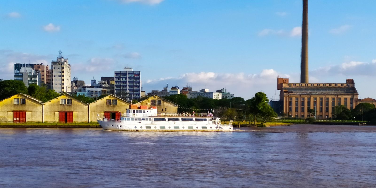 Prédio da antiga termelétrica e sua chaminé altíssima vistos do Guaíba, em Porto Alegre, com um barco no rio